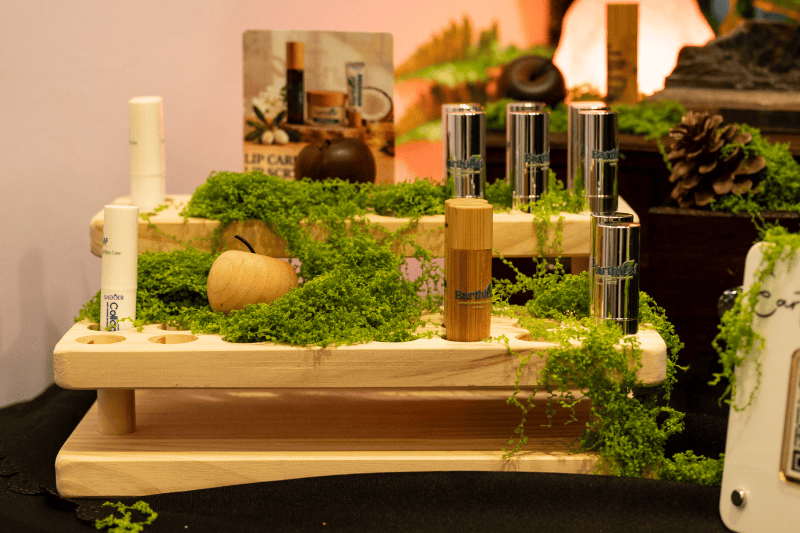 Display of natural lip care products and organic skincare innovations at The Good Life Show Africa event, featuring Earthy branded lip balms in bamboo and metallic tubes arranged on wooden stands with green moss decorations.